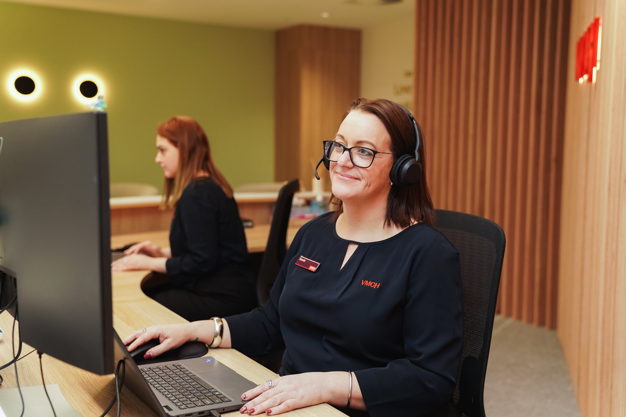 A woman with a headset sitting a reception desk with another woman sitting in the background.