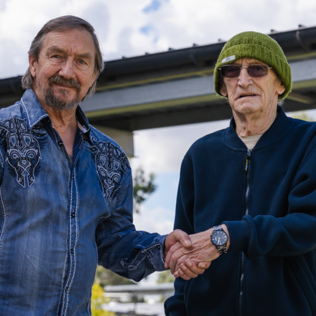 Two VMCH aged care residents smiling and shaking hands outdoors, highlighting friendship and social connection in the community.