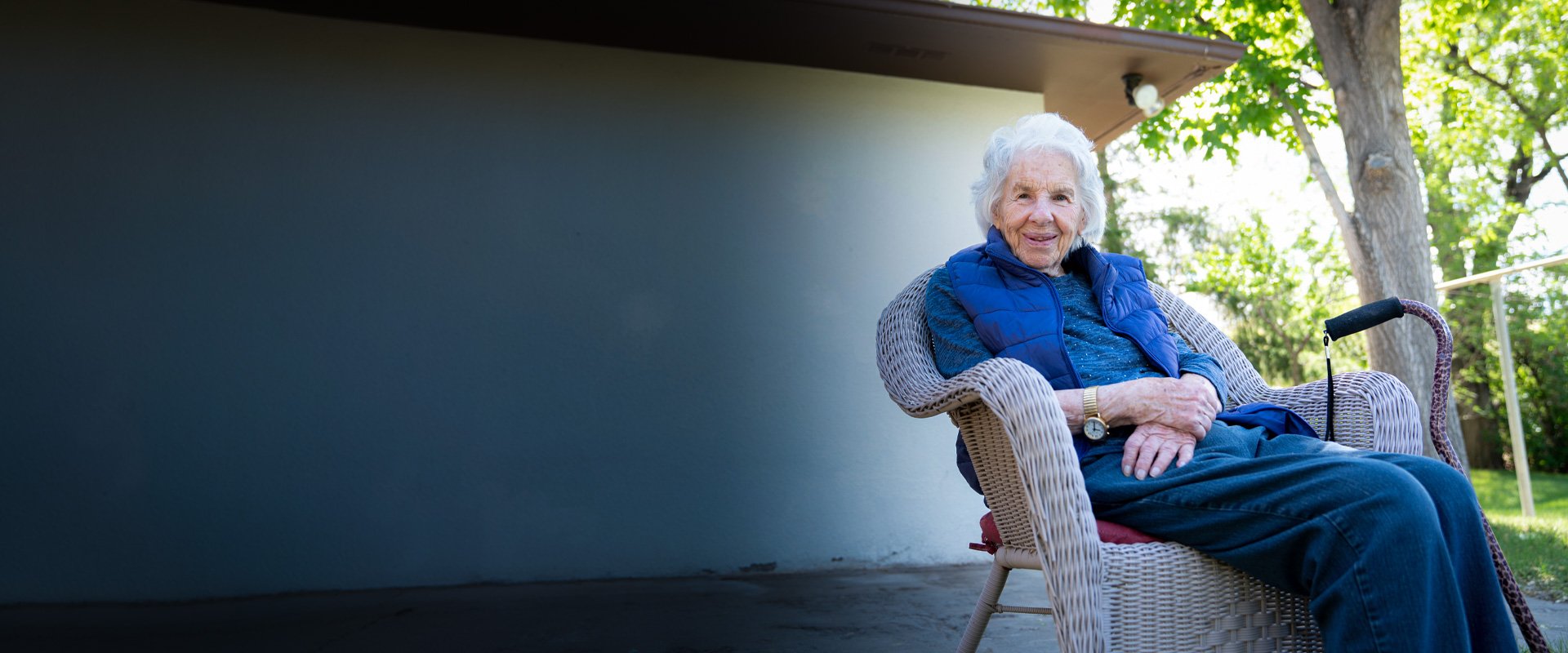 Elderly woman with white hair sits in a wicker chair outdoors, smiling warmly, with a walking stick resting beside her.