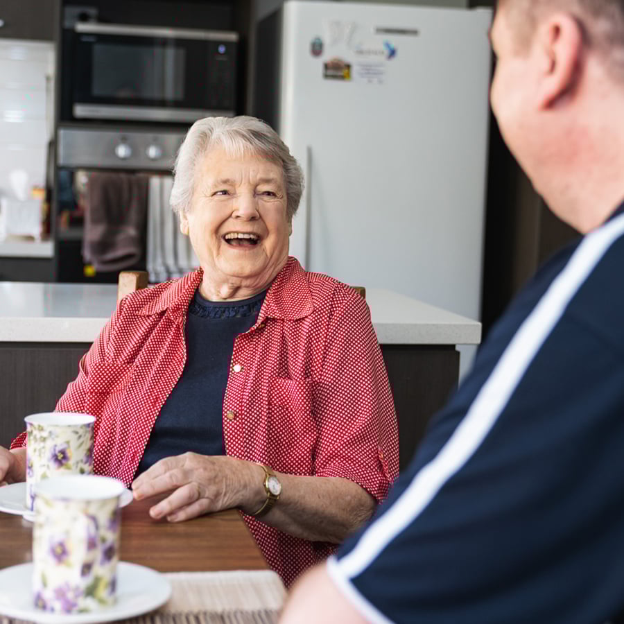 VMCH aged care resident smiling and chatting over tea with a staff member in the kitchen, enjoying companionship and conversation.