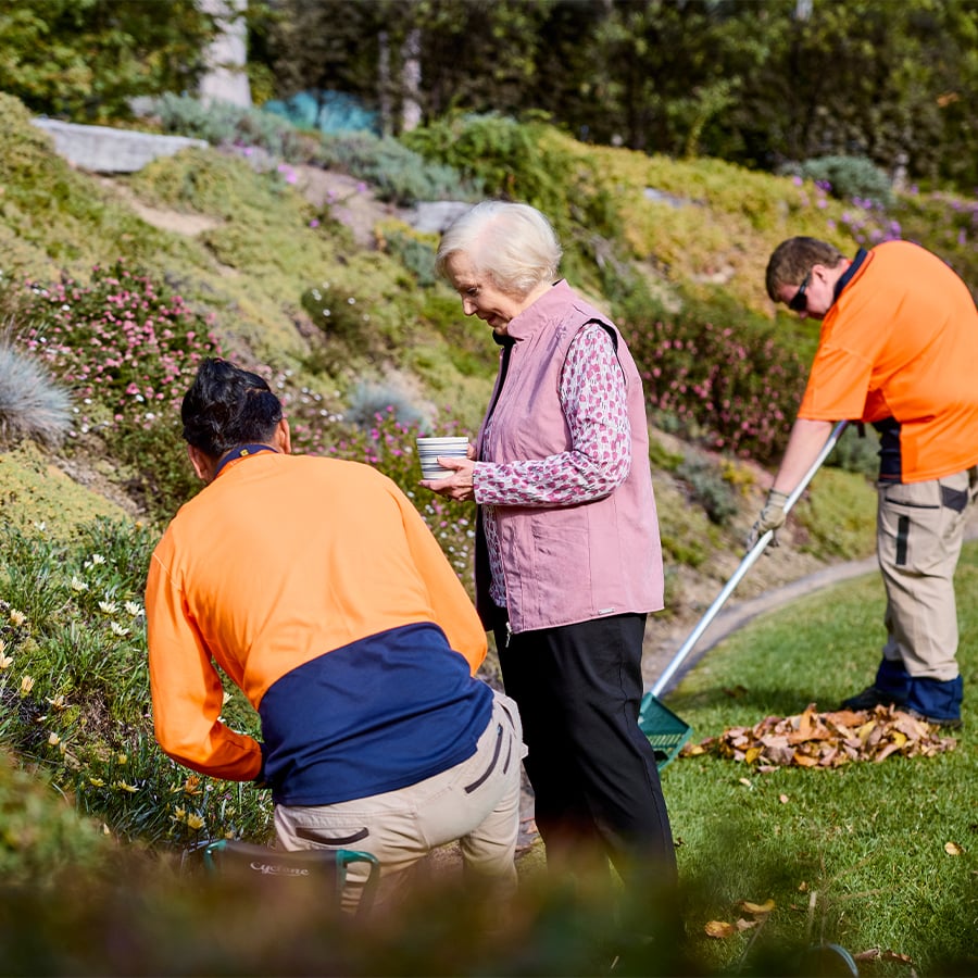 VMCH aged care resident enjoying time outdoors with gardeners, surrounded by greenery and flowers while staff tidy the garden.