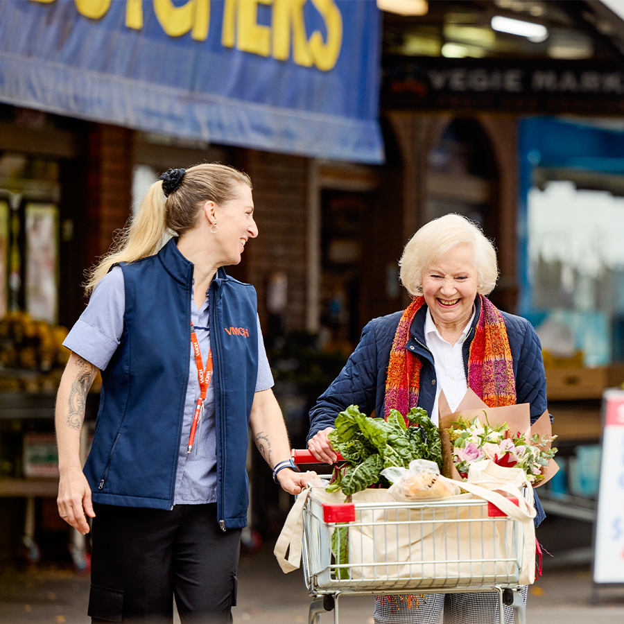 VMCH aged care staff member supporting a resident with grocery shopping, smiling together while pushing a trolley filled with fresh food and flowers.