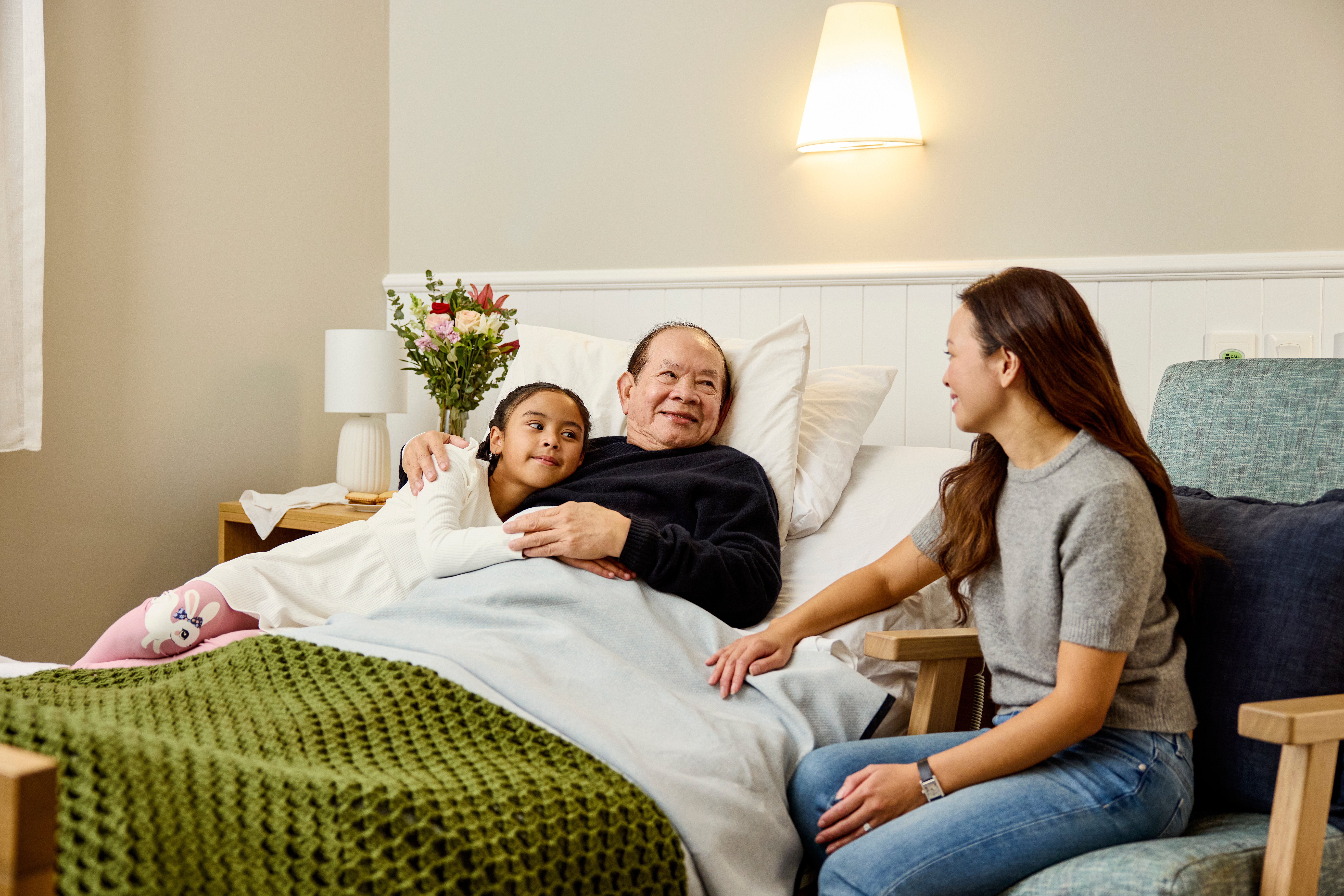 An elderly man in his aged care bed with his granddaughter to his left and daughter to his right. They are smiling.