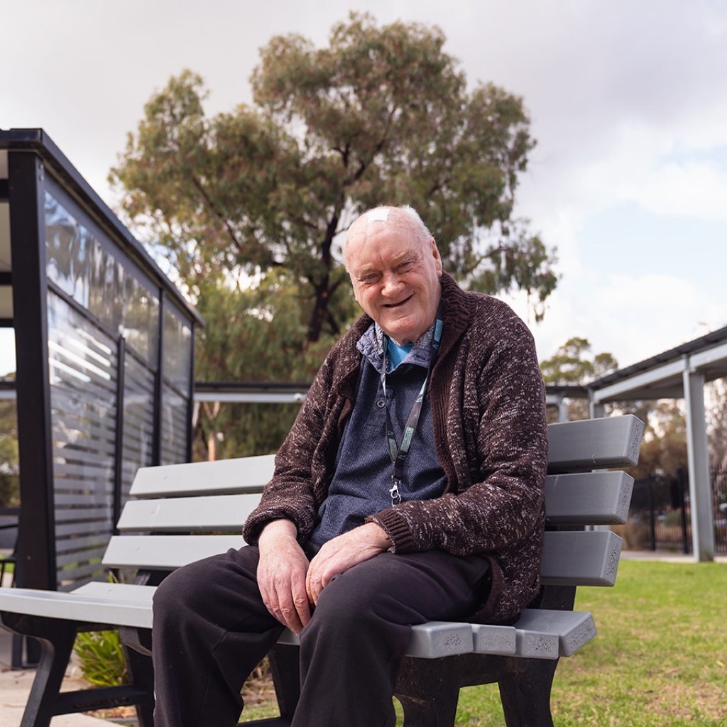 VMCH aged care resident sitting on a bench in the garden, smiling warmly and enjoying the outdoor space.