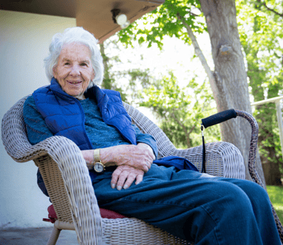 Elderly woman with white hair sits in a wicker chair outdoors, smiling warmly, with a walking stick resting beside her.