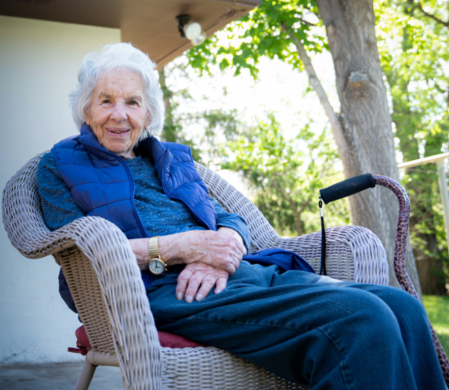 Elderly woman with white hair sits in a wicker chair outdoors, smiling warmly, with a walking stick resting beside her.