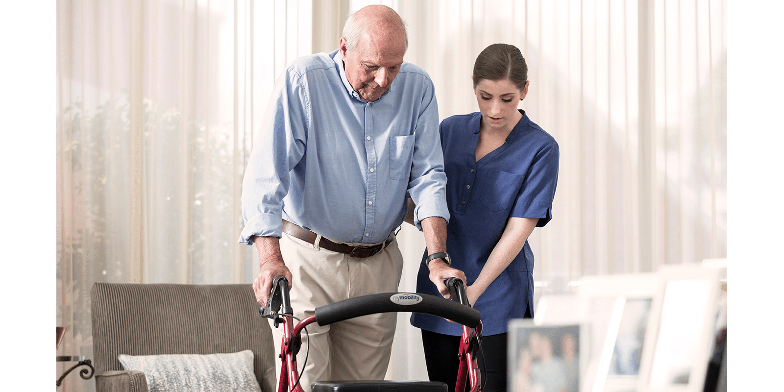 Young female carer assists an elderly man using a red walker in a bright, softly lit living room with sheer curtains and framed photos.