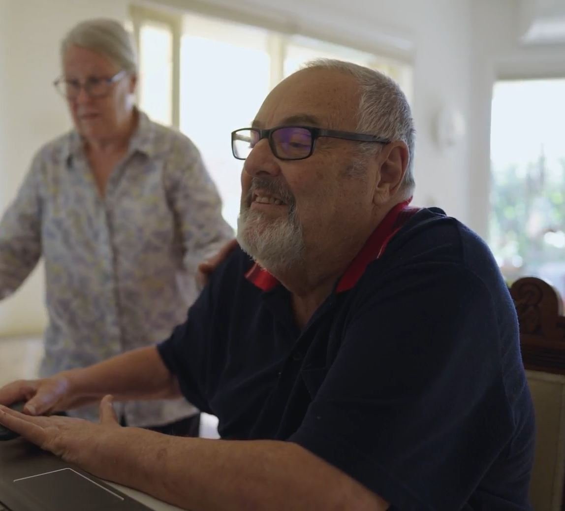 A man sitting at a desk using a computer with a woman walking towards him.